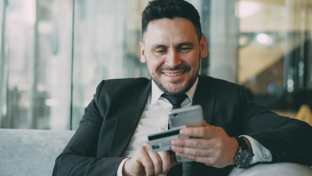 services-05 Cheerful Caucasian businessman in suit using online banking holding credit card and smartphone in his hands in modern cafe indoors during lunch break